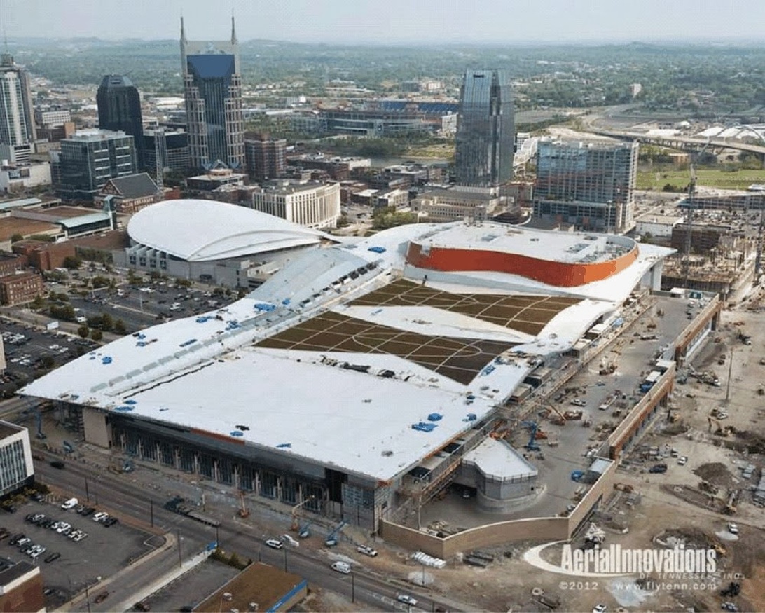 An aerial view of a large building complex under construction in an urban setting, likely a performance venue or convention center with white roof sections and a reddish-orange curved structure. The image shows the building in relation to surrounding city architecture and infrastructure.